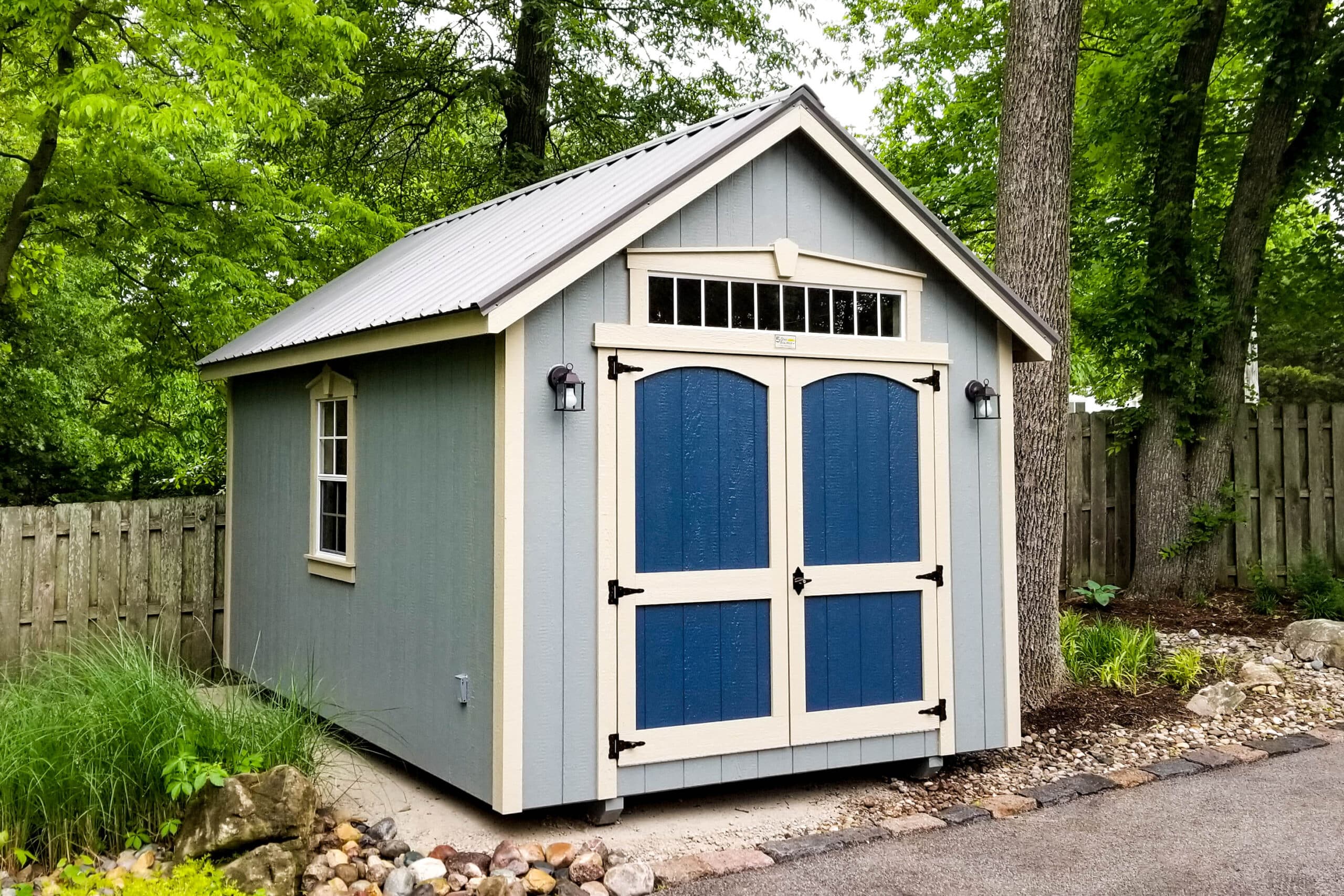 Light blue storage shed with blue double doors, white trim, and a decorative transom window under a gable roof